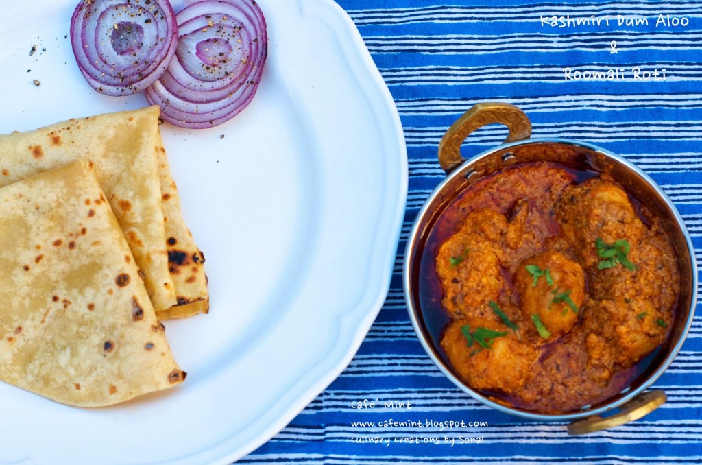 An overhead shot of Kashmir dum aloo in a copper vessel and roomali roti with onion roundels on a white plate on the side with a blue lined backdrop