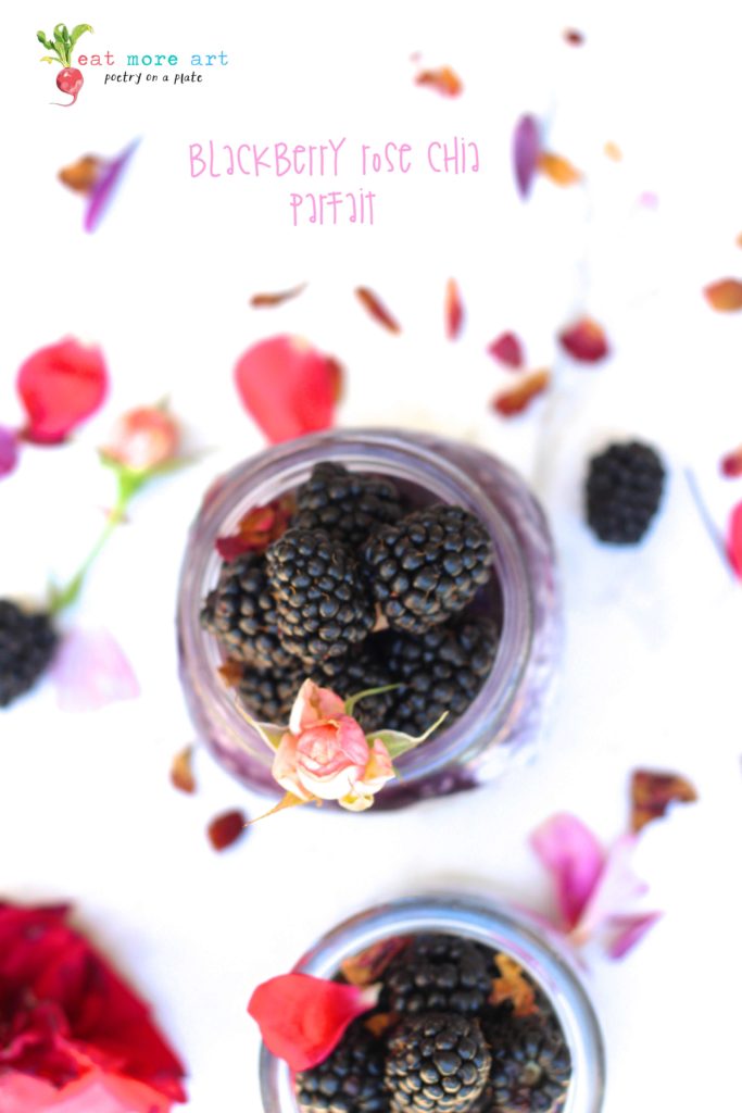 an overhead shot of blackberry rose chia parfait topped with blackberries and rose petals and rose bud on a white backdrop with flower petals and blueberries scattered